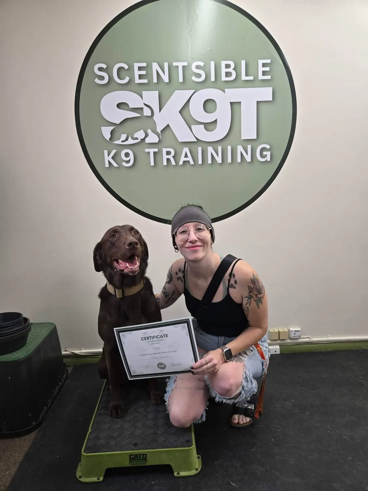 A chocolate lab and their handler holding a graduation certificate in front of the Scentsible K9 logo wall