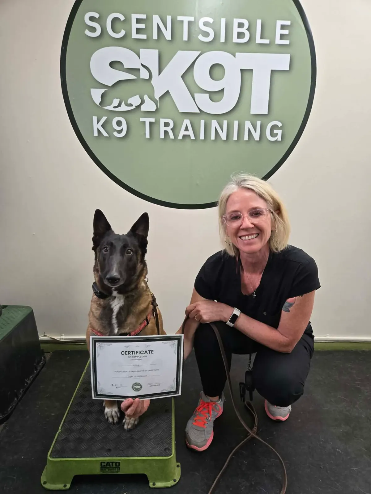 A Belgian malinois sitting square on the platform beside their handler and a graduation certificate