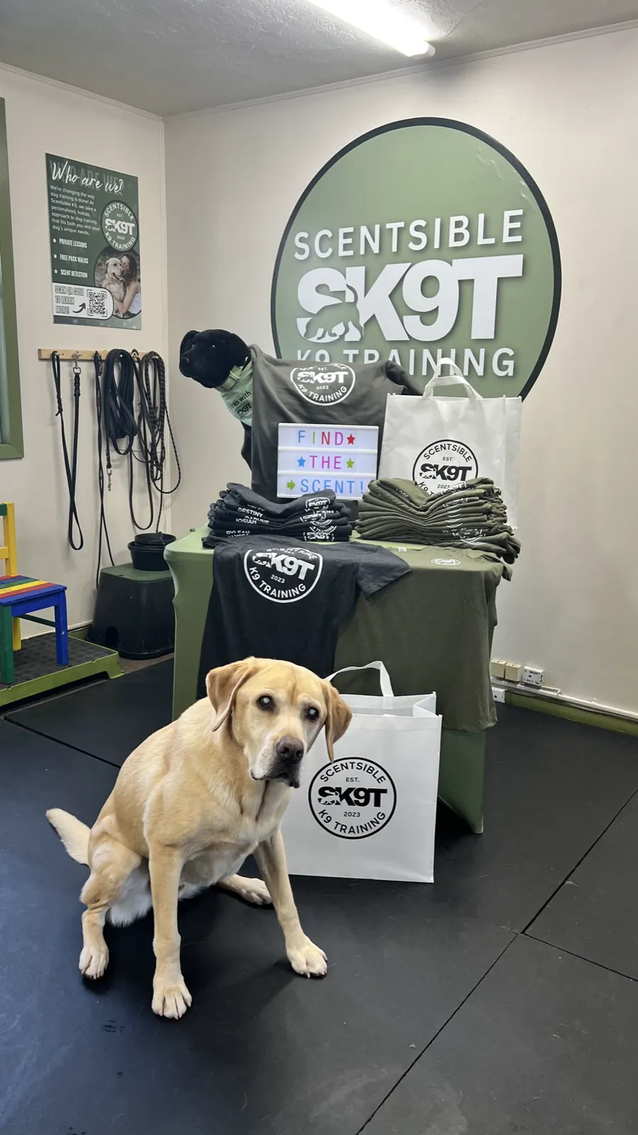 A yellow lab sitting beside a Scentsible K9 tote bag at the merch table